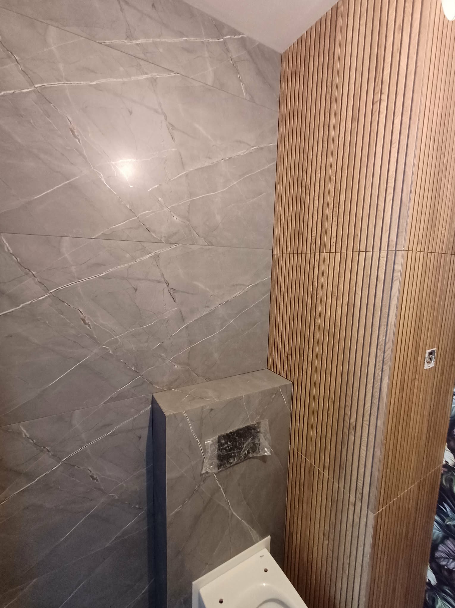 Wykończona łazienka z szarymi płytkami imitującymi marmur i drewnianą ścianą z pionowych paneli. Widoczny fragment toalety i zabudowa stelaża podtynkowego.