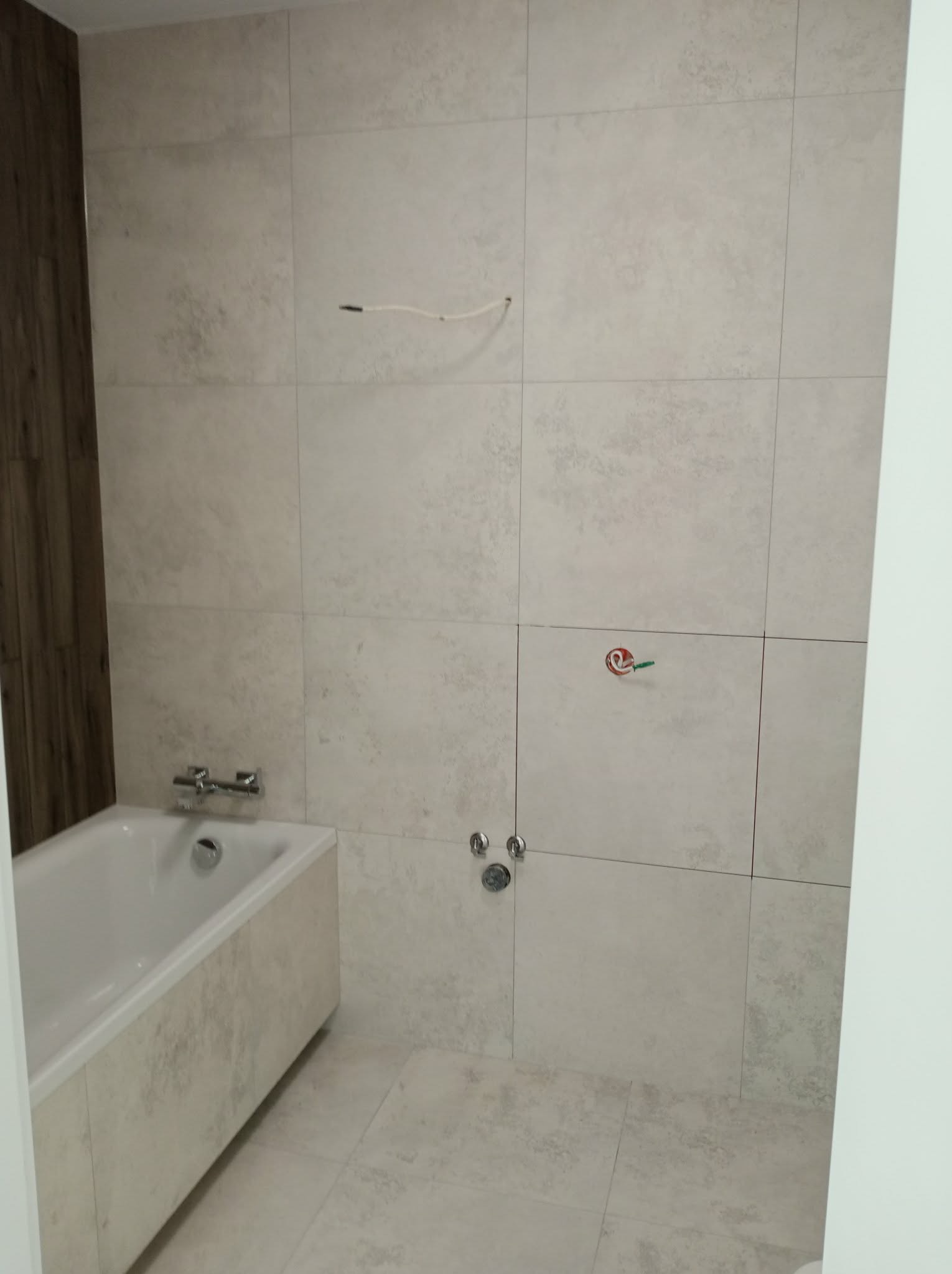 Wykończona łazienka z wanną obłożoną płytkami w odcieniach beżu i szarości. Na ścianie widoczne przyłącza wodne i fragment drewnianej okładziny.