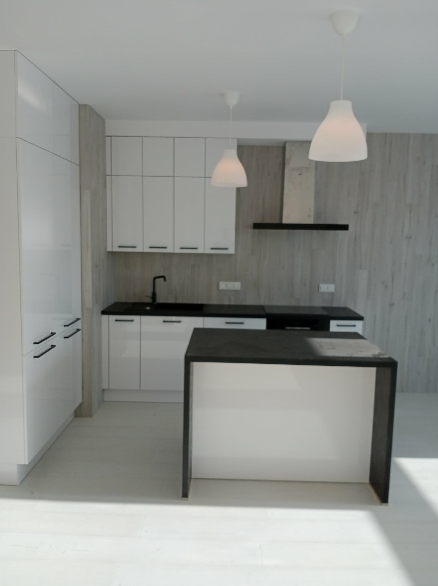 Nowoczesna, minimalistyczna kuchnia z białymi szafkami, czarnym blatem i wyspą. Trzy lampy wiszące nad wyspą. Ściana wykończona panelami imitującymi drewno.
