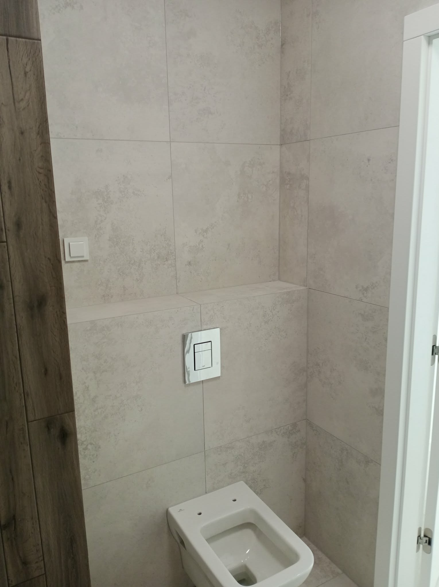 Wykończona łazienka z beżowymi płytkami imitującymi kamień, podwieszana toaleta z chromowanym przyciskiem, drewnopodobna obudowa i białe drzwi. Minimalistyczny design.