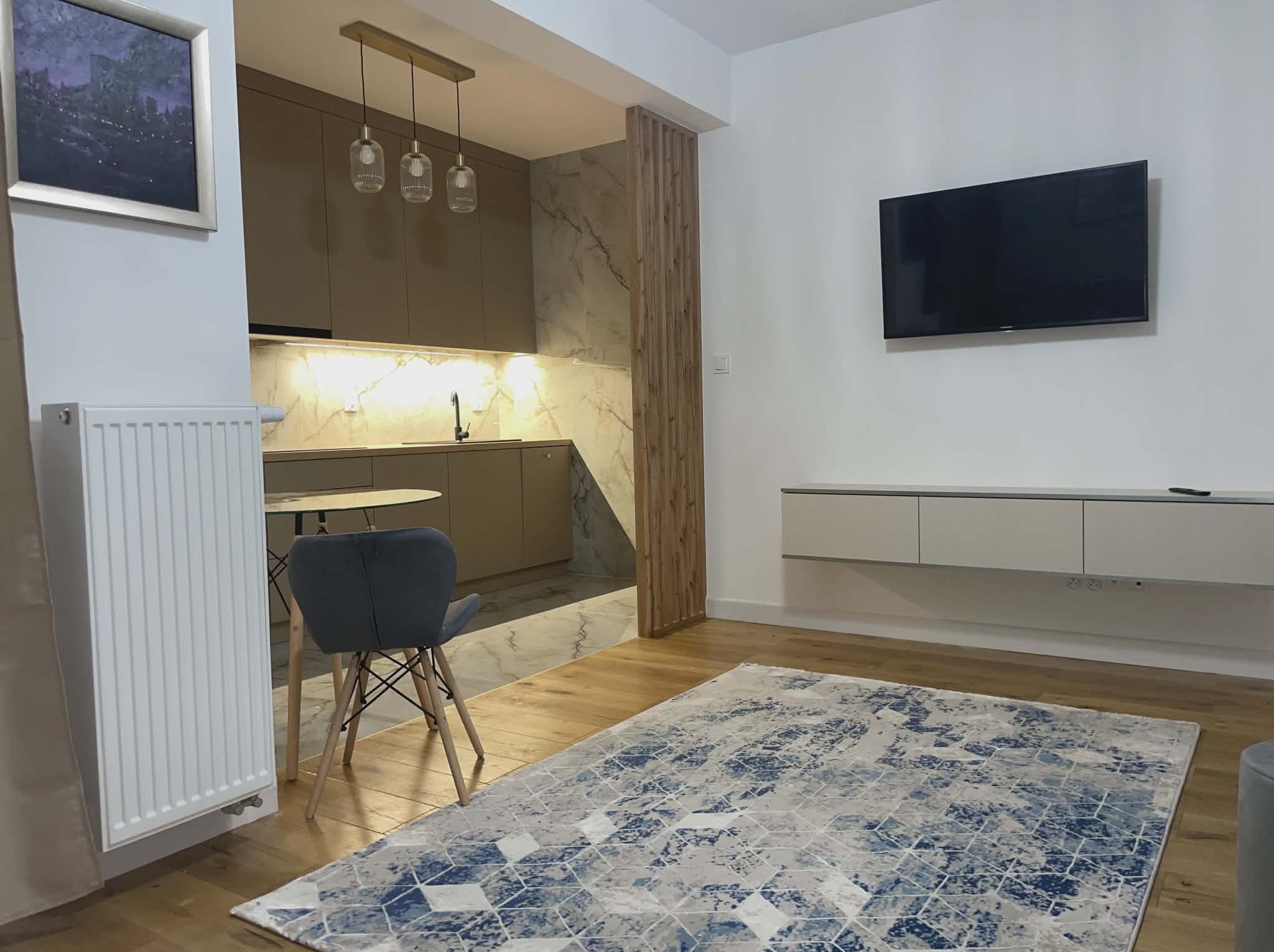 Nowoczesne wnętrze: beżowa kuchnia z marmurowym blatem, drewniana ścianka działowa, TV na ścianie, dywan w geometryczne wzory, krzesło i grzejnik.