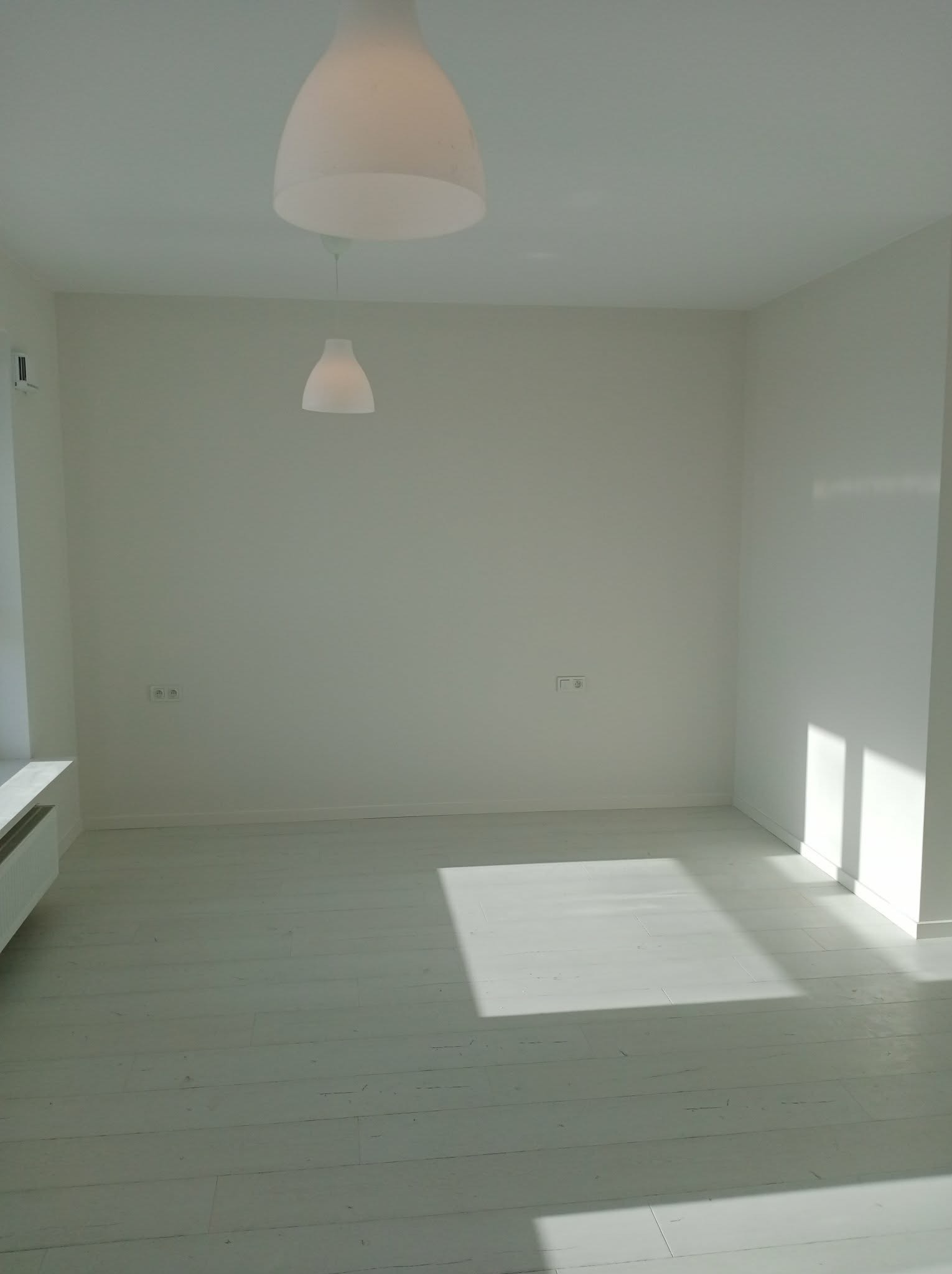 Jasne, minimalistyczne wnętrze po remoncie: białe ściany, podłoga z desek, nowoczesne lampy wiszące. Wpadające promienie słoneczne podkreślają czystość i świeżość.