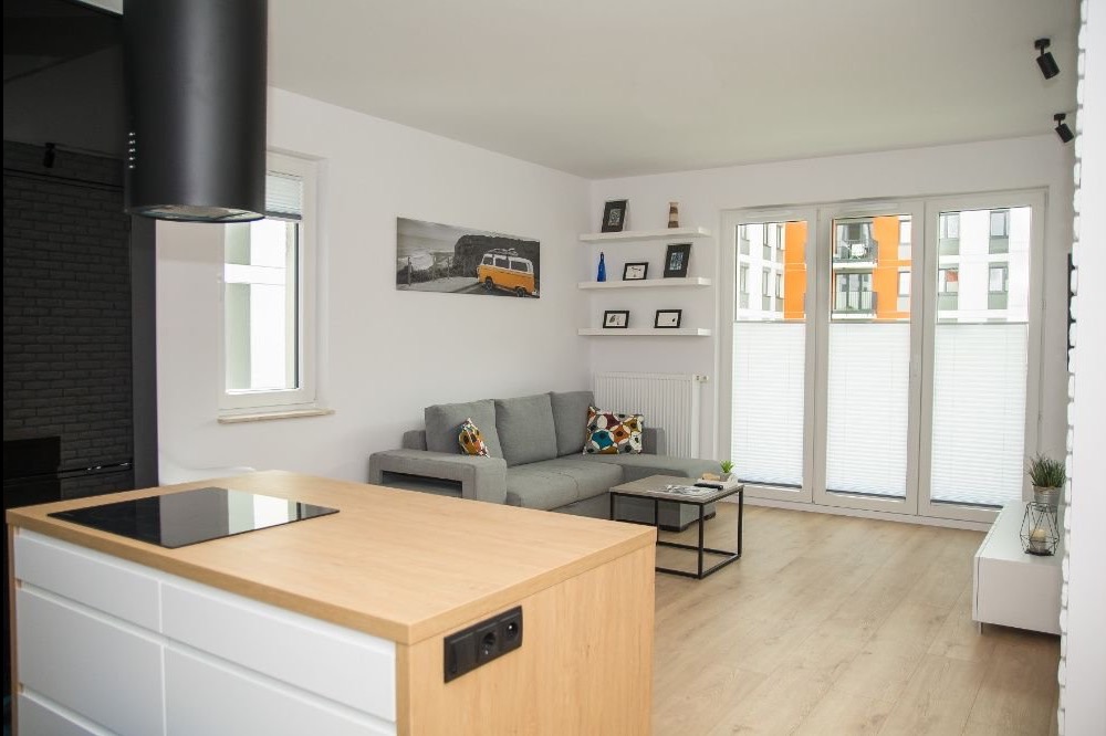 Jasne wnętrze salonu z aneksem kuchennym: szara sofa, drewniany parkiet, białe ściany, okno z roletami plisowanymi i wyspa kuchenna z płytą indukcyjną.