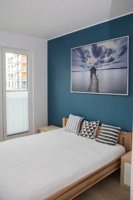 Błękitna ściana z obrazem nad łóżkiem w jasnej sypialni. Widok na okno z żaluzjami i budynek w tle. Minimalistyczny design wnętrza.
