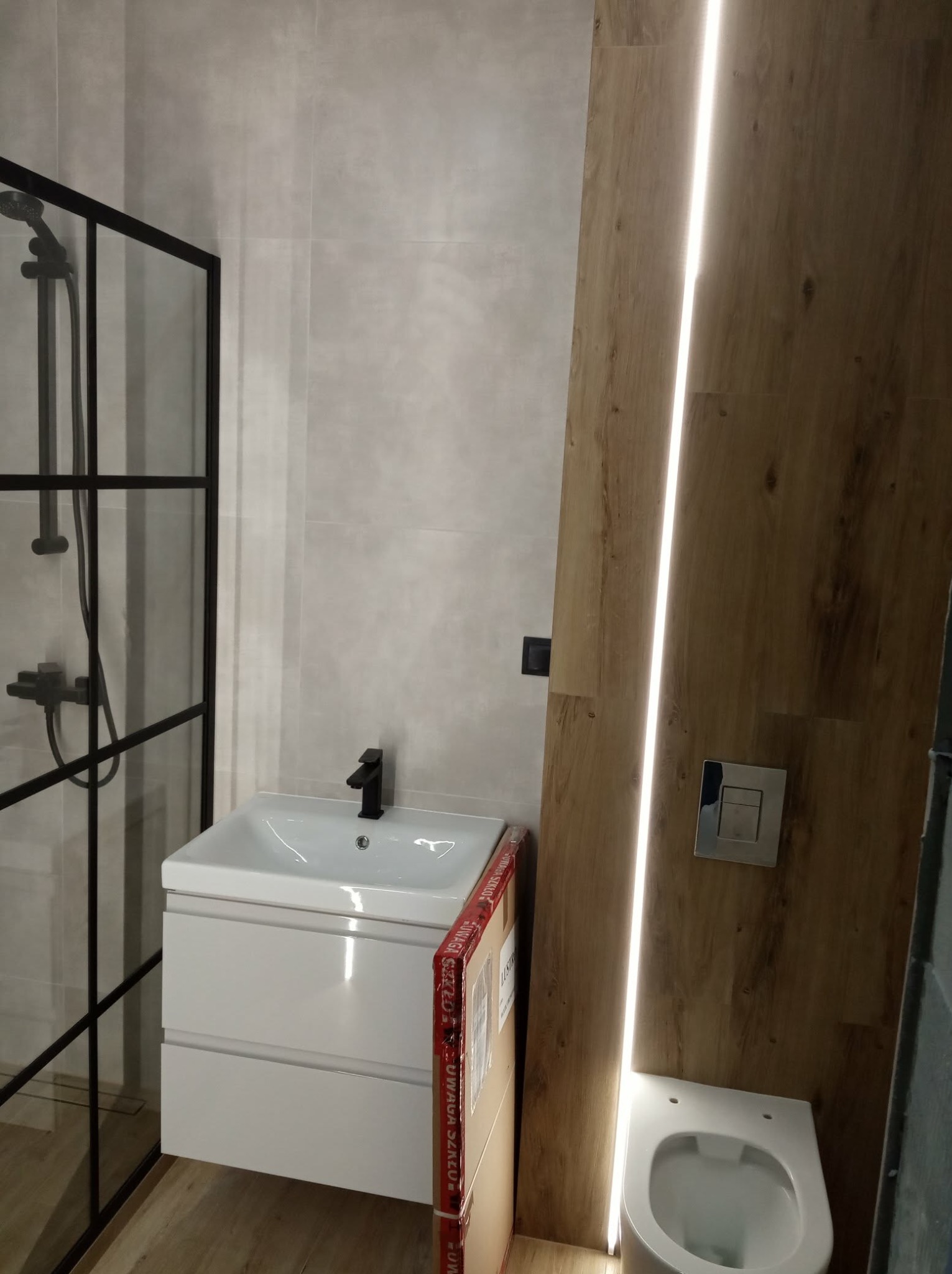 Nowoczesna łazienka z kabiną prysznicową typu walk-in, białą szafką z umywalką i podświetlaną drewnopodobną ścianą. Minimalistyczny design, czarne akcenty.