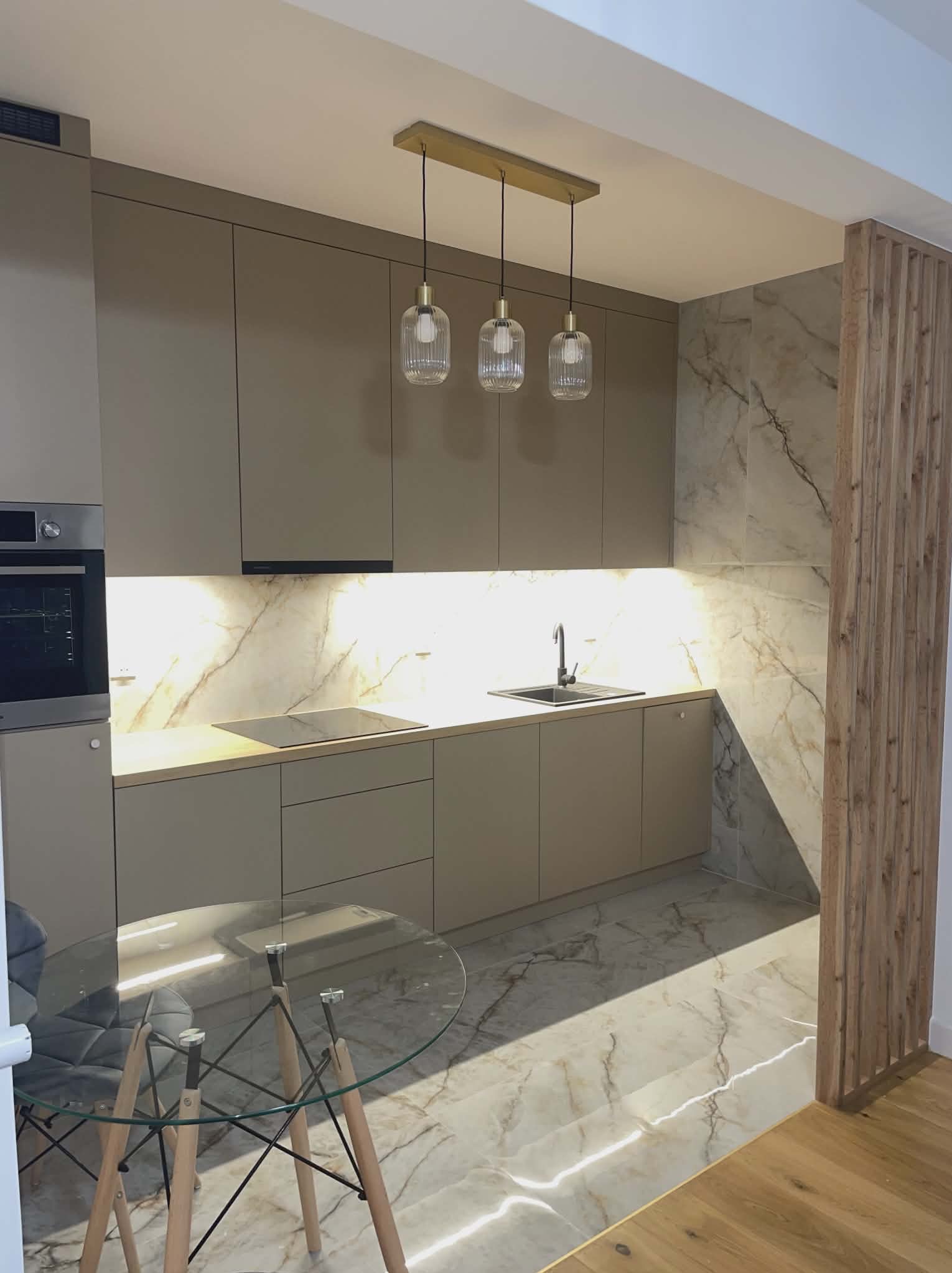 Nowoczesna kuchnia w odcieniach beżu z marmurowym wykończeniem ścian i podłogi. Szklany stół z krzesłami w tle. Oświetlenie punktowe nad blatem.