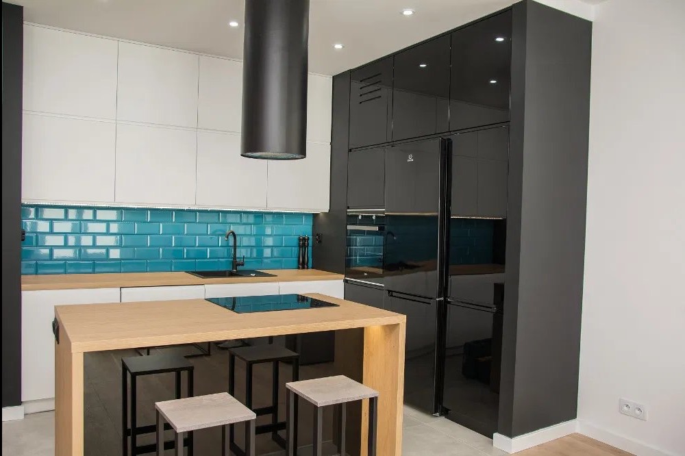 Nowoczesna kuchnia z wyspą: białe szafki, turkusowe płytki, czarny okap cylindryczny i zabudowa z czarnym połyskiem, minimalistyczne stołki barowe.