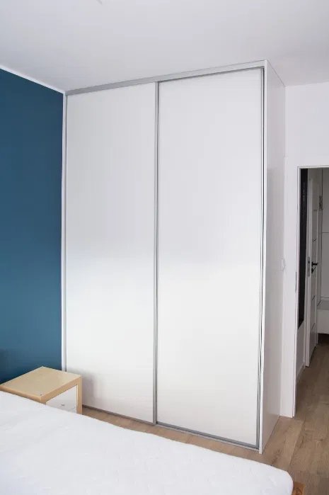 Biała szafa przesuwna z srebrnymi obramowaniami w sypialni z niebieską ścianą. Widoczny fragment łóżka i szafki nocnej. Minimalistyczny design.