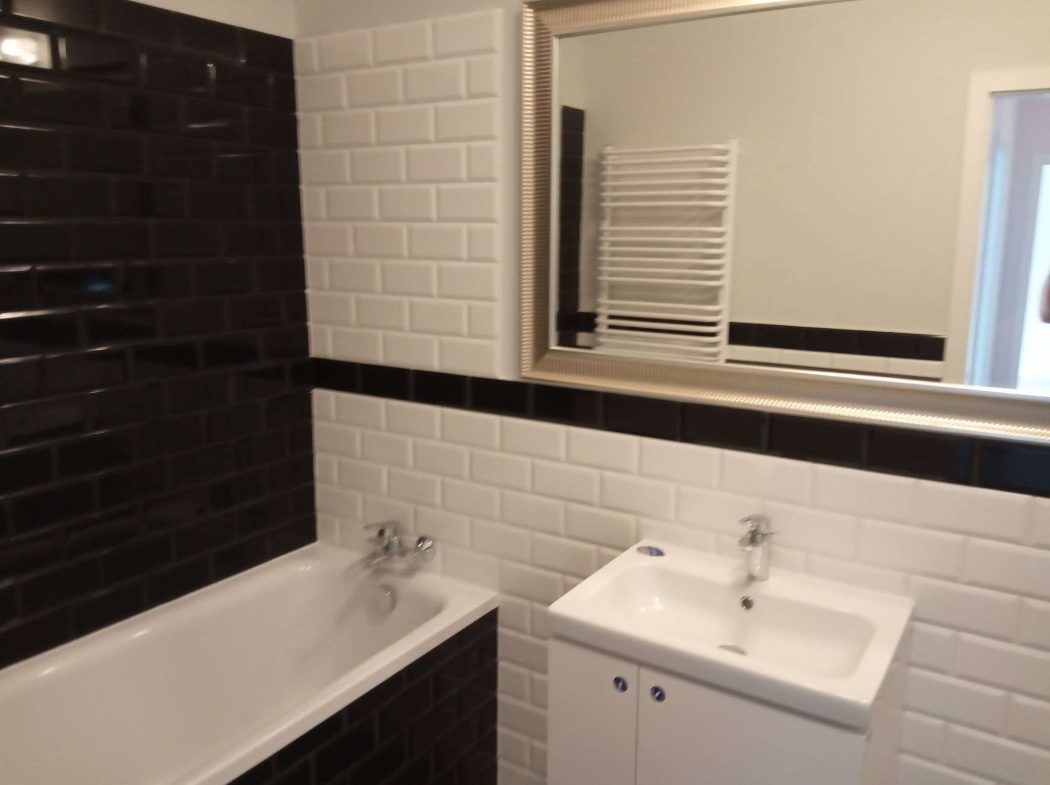 Biało-czarna łazienka z wanną i umywalką, wykończona płytkami typu metro. W lustrze odbija się grzejnik drabinkowy. Kontrastowe barwy i nowoczesny design.