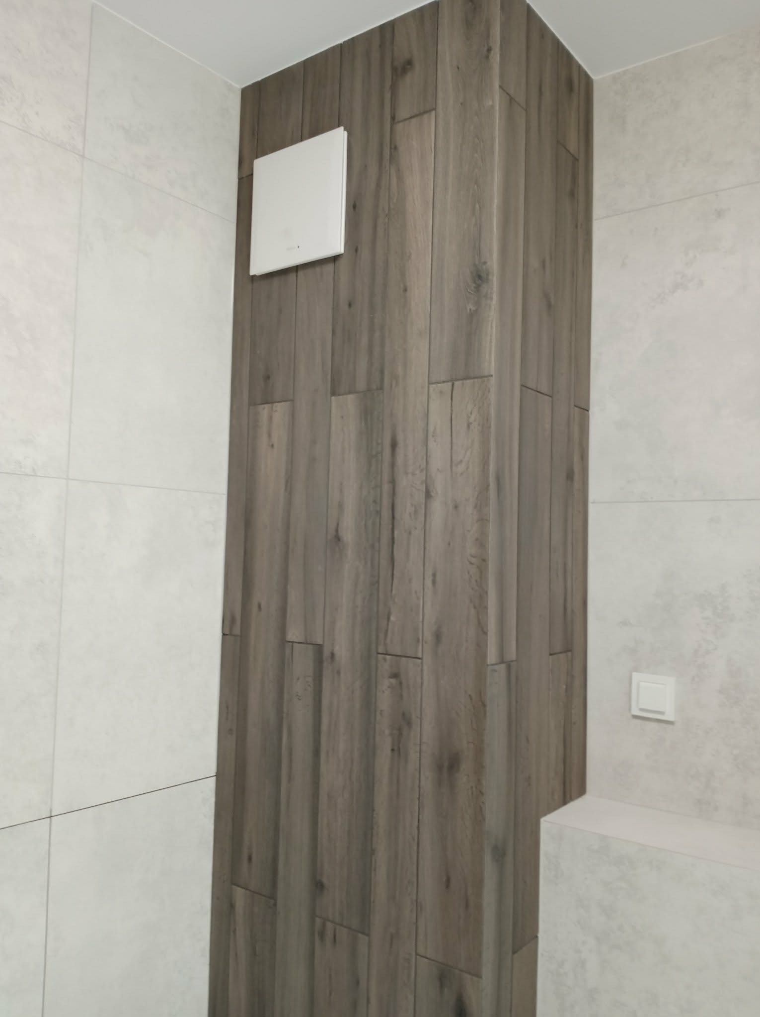 Wykończenie ściany w łazience w stylu drewnianych paneli, z kratką wentylacyjną i włącznikiem światła. Realizacja z Warszawy.