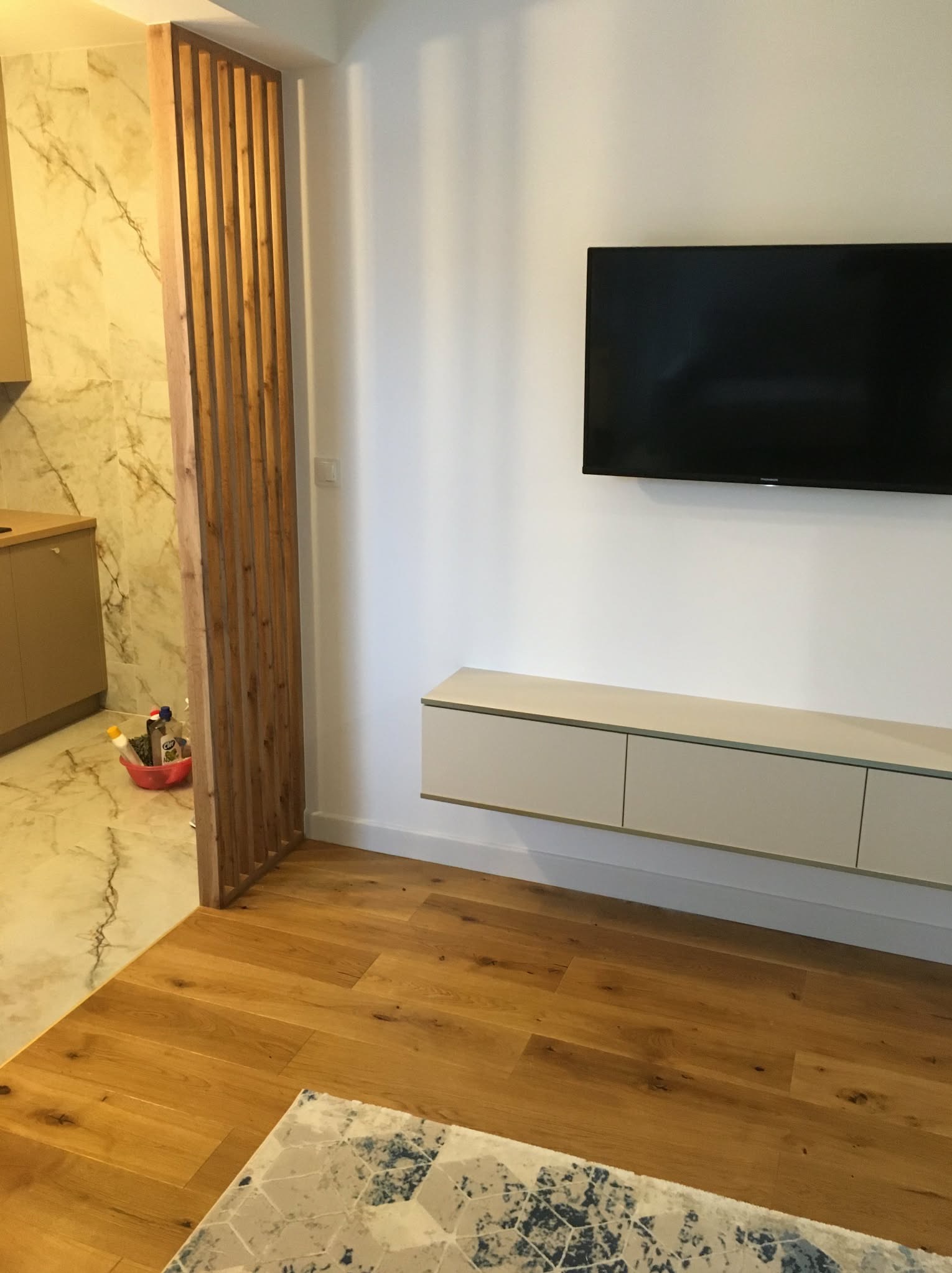 Nowoczesne wnętrze: drewniana podłoga, białe ściany z TV, beżowa szafka RTV i ażurowa ścianka działowa oddzielająca kuchnię z marmurowymi płytkami.