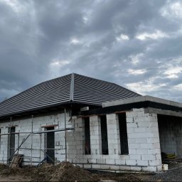 Mateusz Filipczak TechBud BGT - Dom w trakcie budowy z ułożonym, ciemnym dachem z dachówki ceramicznej. Widoczne białe bloczki konstrukcyjne i rusztowania. Pochmurne niebo w tle.