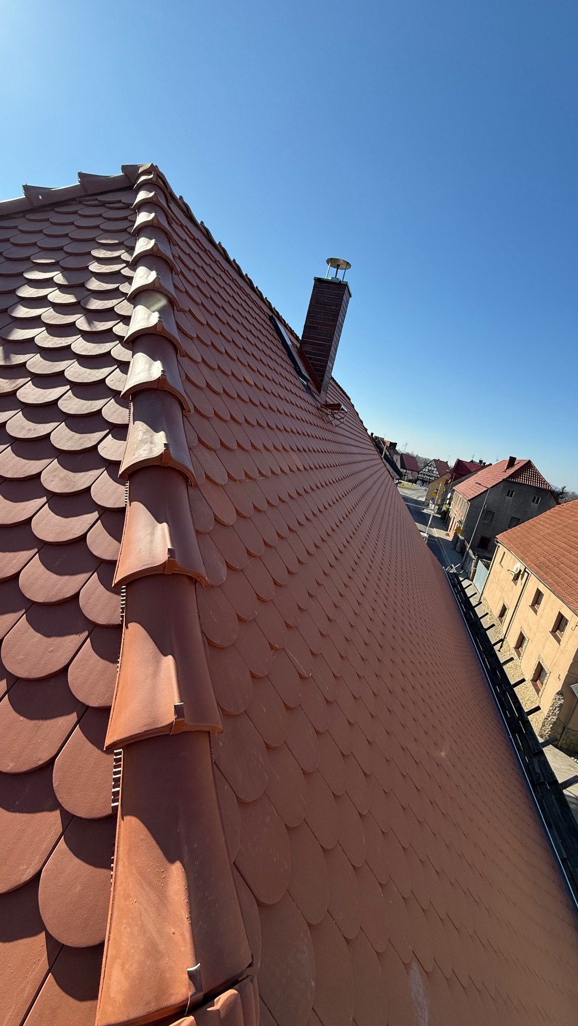 Czerwony dach z dachówki karpiówki, widok z góry, komin z cegły, błękitne niebo w tle, budynki mieszkalne w oddali. Dach po renowacji.