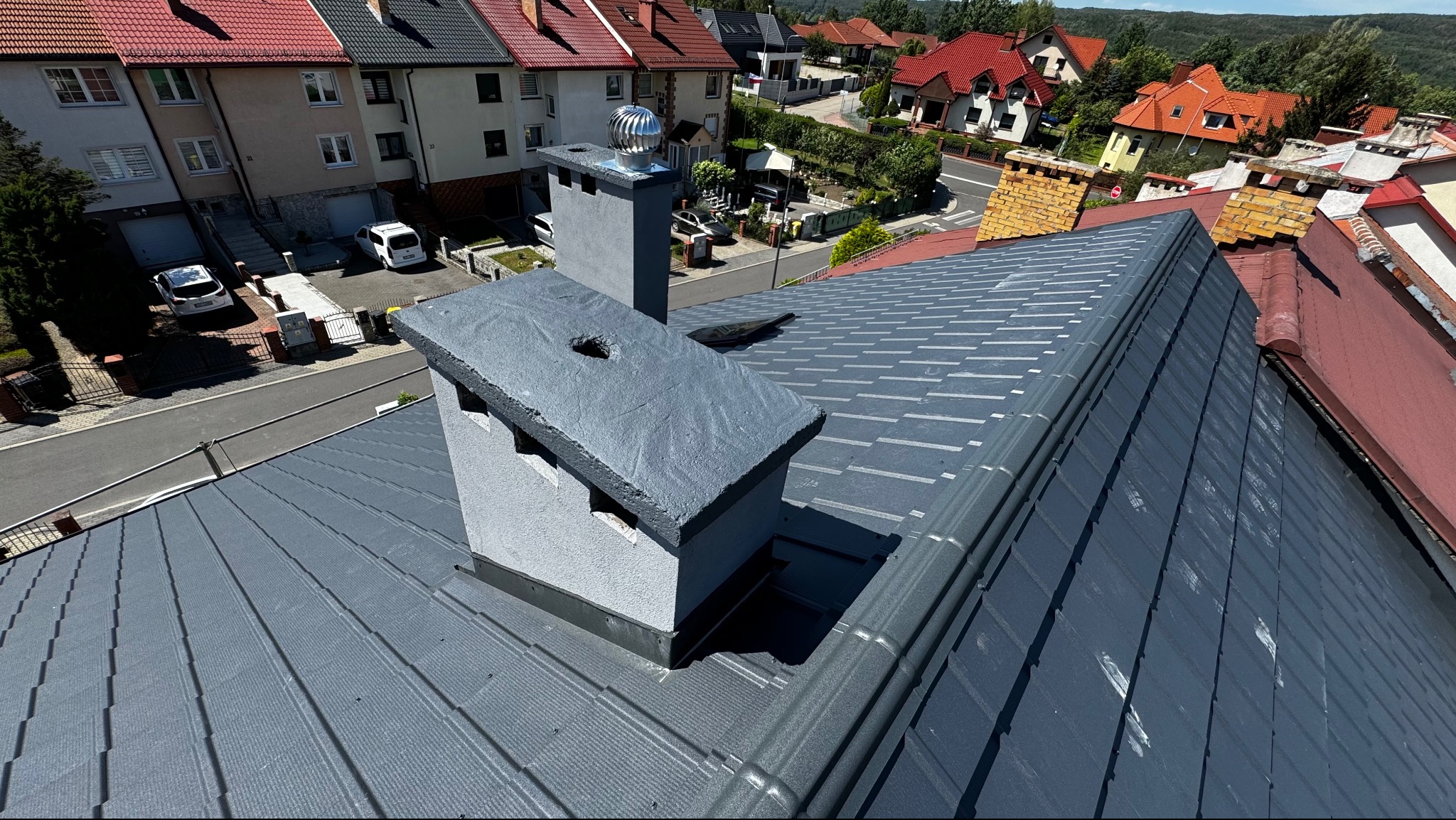 Szary dach z blachy trapezowej z kominem i turbiną w tle zabudowy miejskiej. Widok z góry.