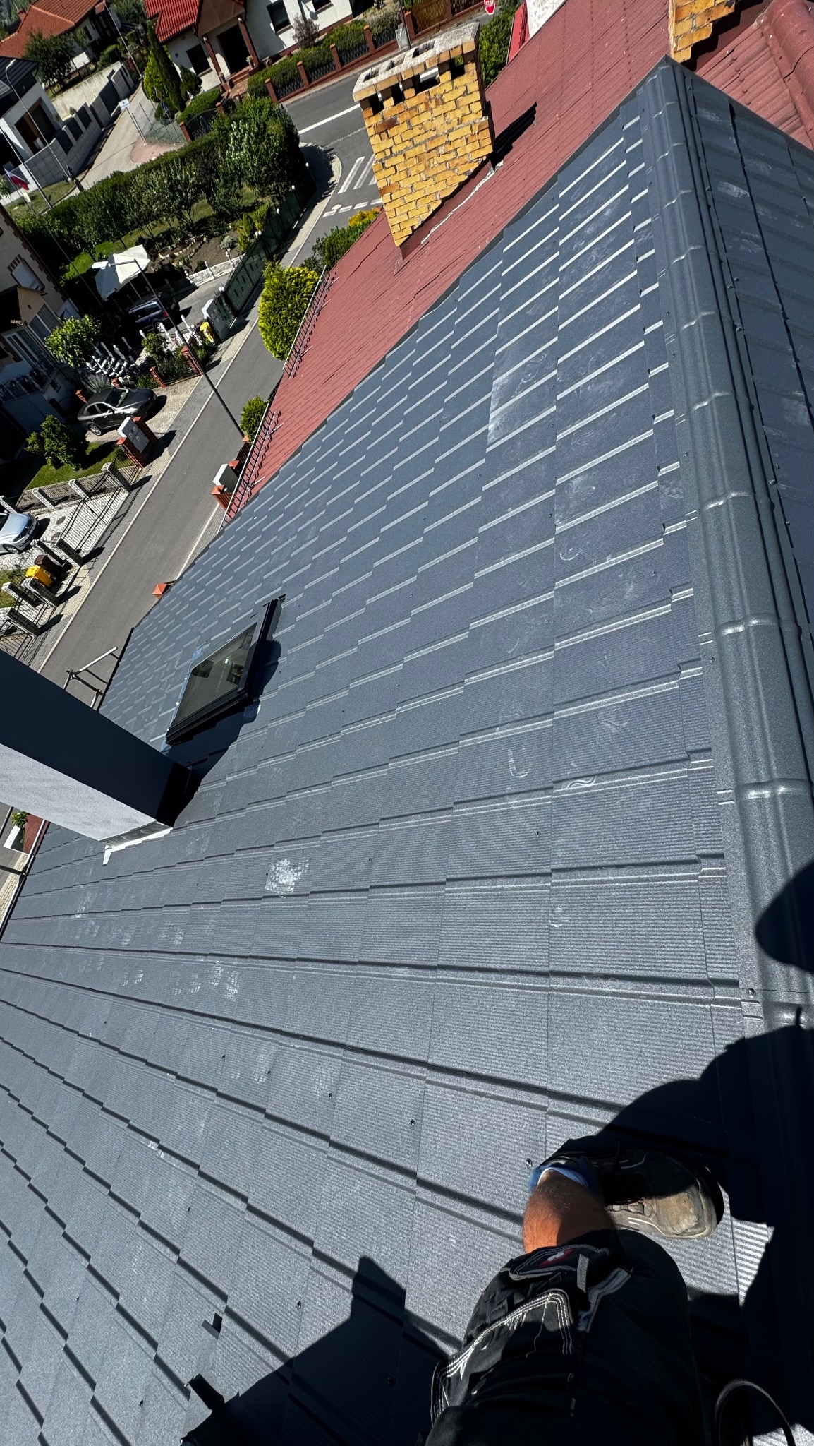 Szary dach z blachodachówki z oknem dachowym, widok z góry, w tle domy i ulica. Fragment sylwetki dekarza w prawym dolnym rogu.