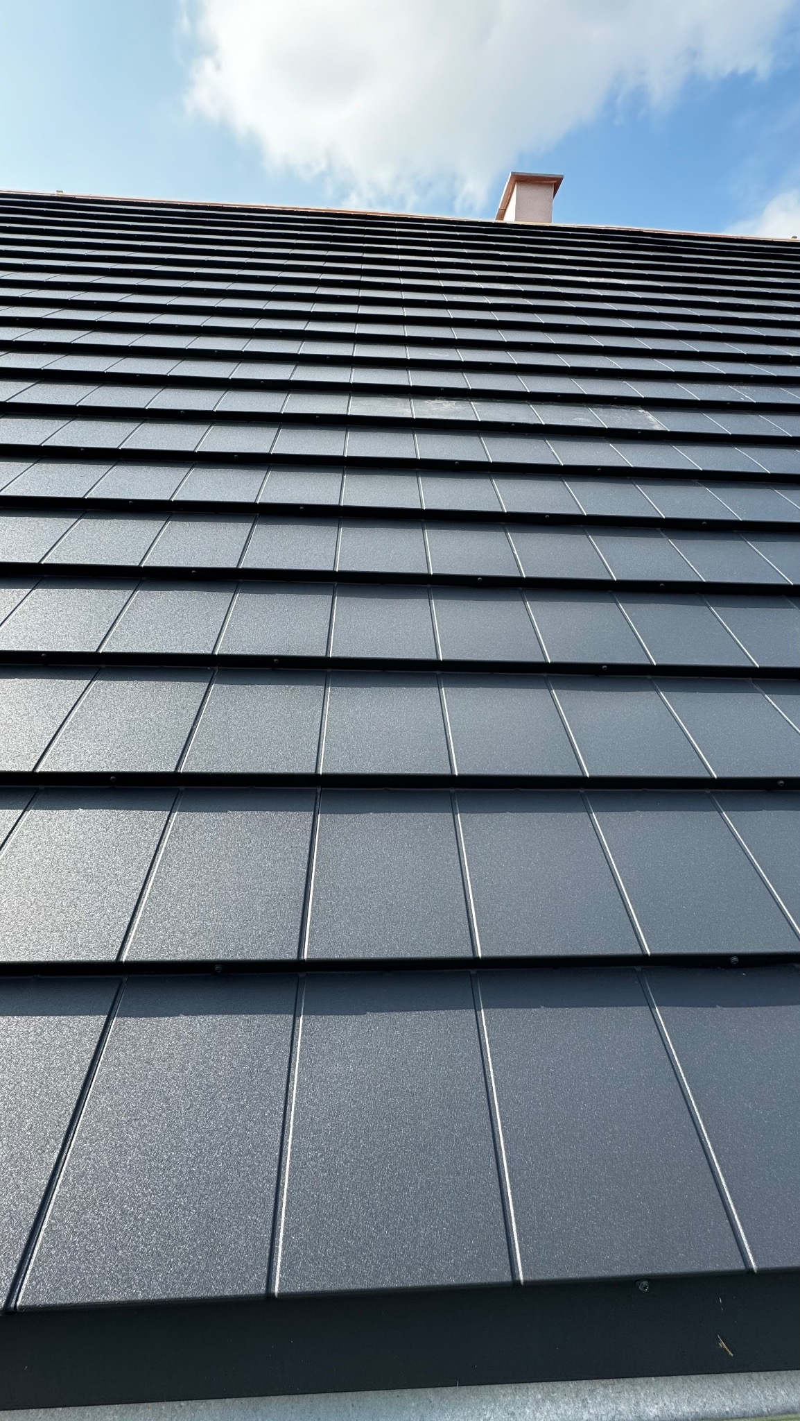 Szczegółowe ujęcie ciemnoszarego dachu z blachy na tle błękitnego nieba z kominem w oddali, ukazujące regularny wzór paneli i fakturę materiału.