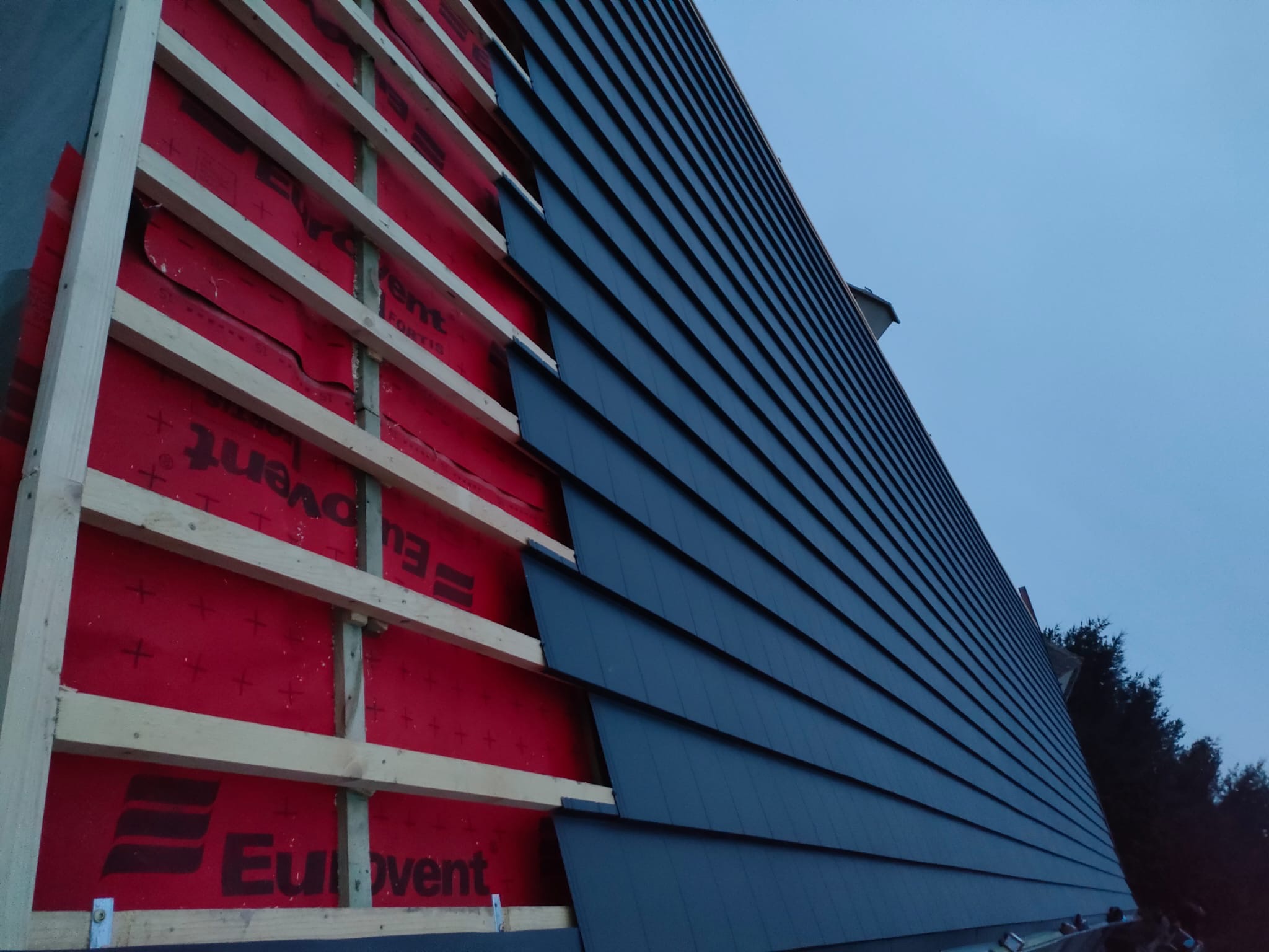 Fragment dachu w trakcie krycia: widoczna więźba, czerwona membrana i ułożona część ciemnych dachówek. Niebo w tle.