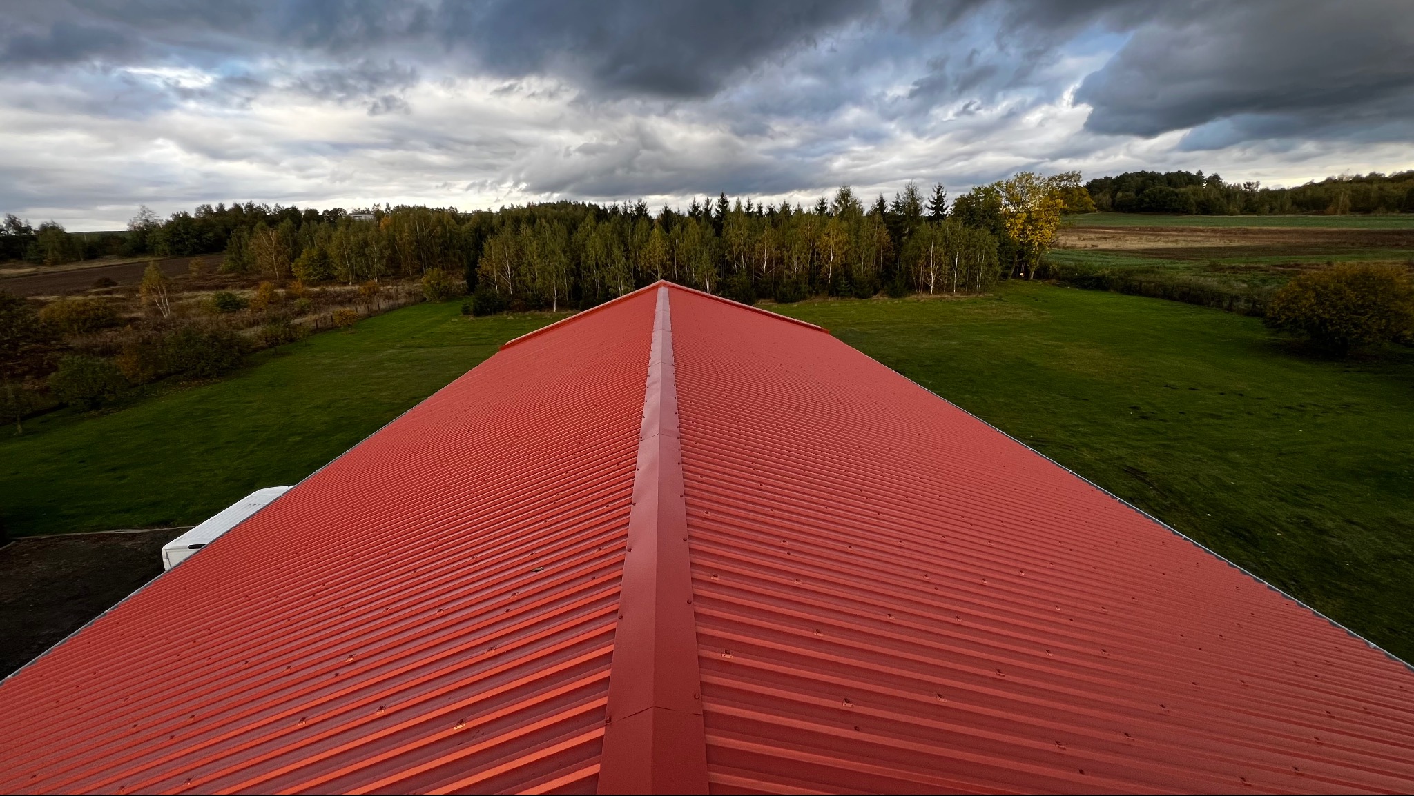 Czerwony, blaszany dach z wyraźnymi żłobieniami, widziany z góry, z zielonym tłem drzew i pochmurnym niebem. Dach jest symetryczny, z wyraźnym grzbietem.