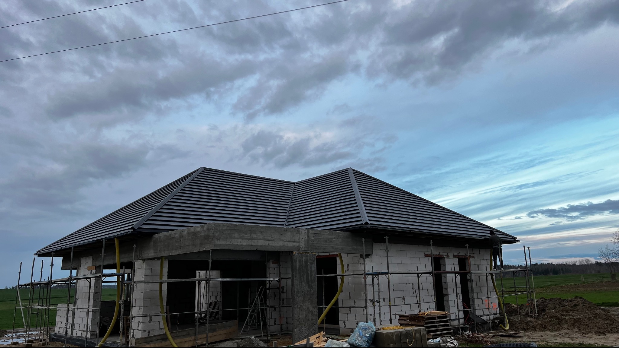 Dom w trakcie budowy z częściowo ukończonym dachem z szarej dachówki, widoczne rusztowania i materiały budowlane na tle pochmurnego nieba.