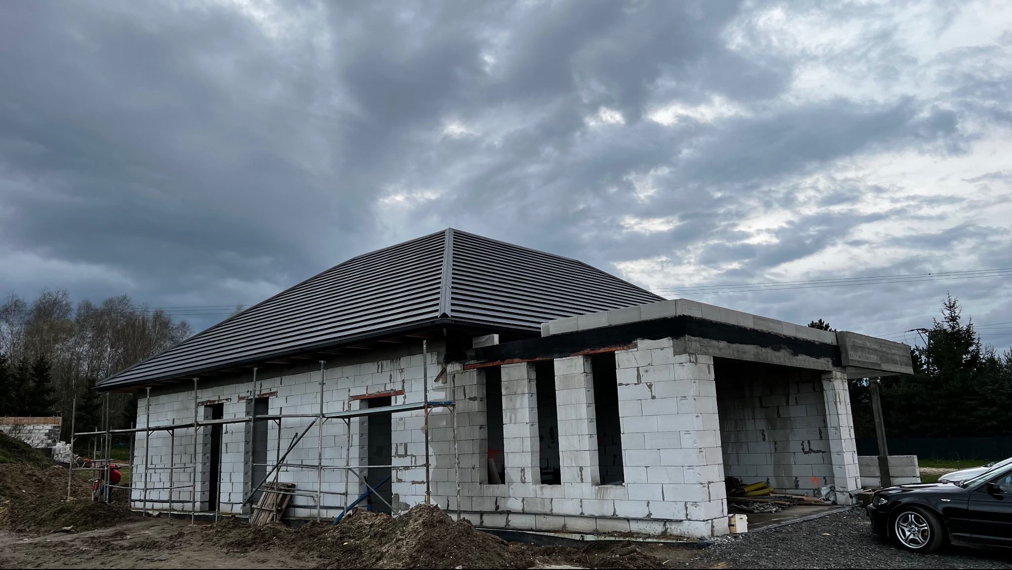 Dom w trakcie budowy z ułożonym, ciemnym dachem z dachówki ceramicznej. Widoczne białe bloczki konstrukcyjne i rusztowania. Pochmurne niebo w tle.