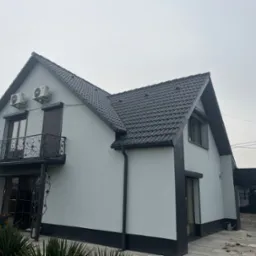 Dom jednorodzinny z ciemną dachówką i jasnoszarą elewacją. Widoczne rynny, okna z roletami i balkon z ozdobną, metalową balustradą. Na elewacji zamontowane klimatyzatory.