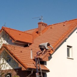 Mirosław Marciniak - Pracownik zabezpieczony linami na dachu z czerwonej dachówki, przy kominie z cegły i drabinie, podczas prac dekarskich w słoneczny dzień.