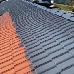 Częściowo pomalowany dach z dachówki ceramicznej, z widocznym podziałem na starą czerwoną i nową szarą farbę. Słoneczny dzień, widok z góry.