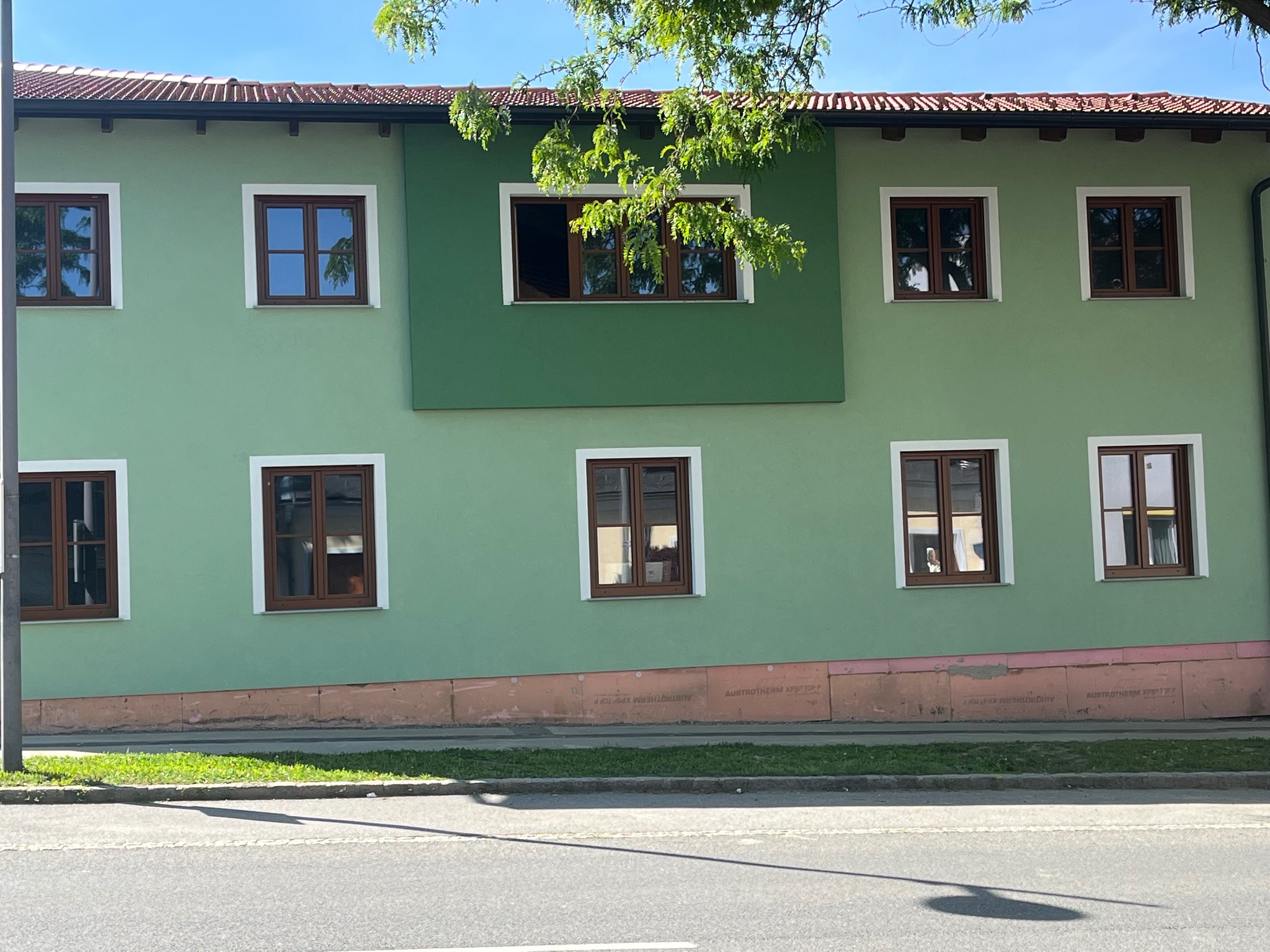 Zielona elewacja dwukondygnacyjnego budynku z brązowymi oknami i białymi obramowaniami. Dolna część elewacji ocieplona płytami. Fragment drzewa w górnej części kadru.