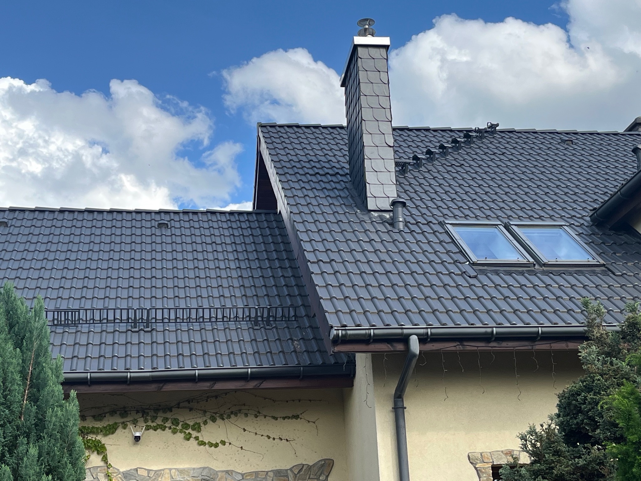 Dom z ciemnoszarą dachówką, widoczne okna dachowe, rynny i komin obłożony łupkiem. Na elewacji widoczny kamień i bluszcz. Błękitne niebo z białymi chmurami w tle.