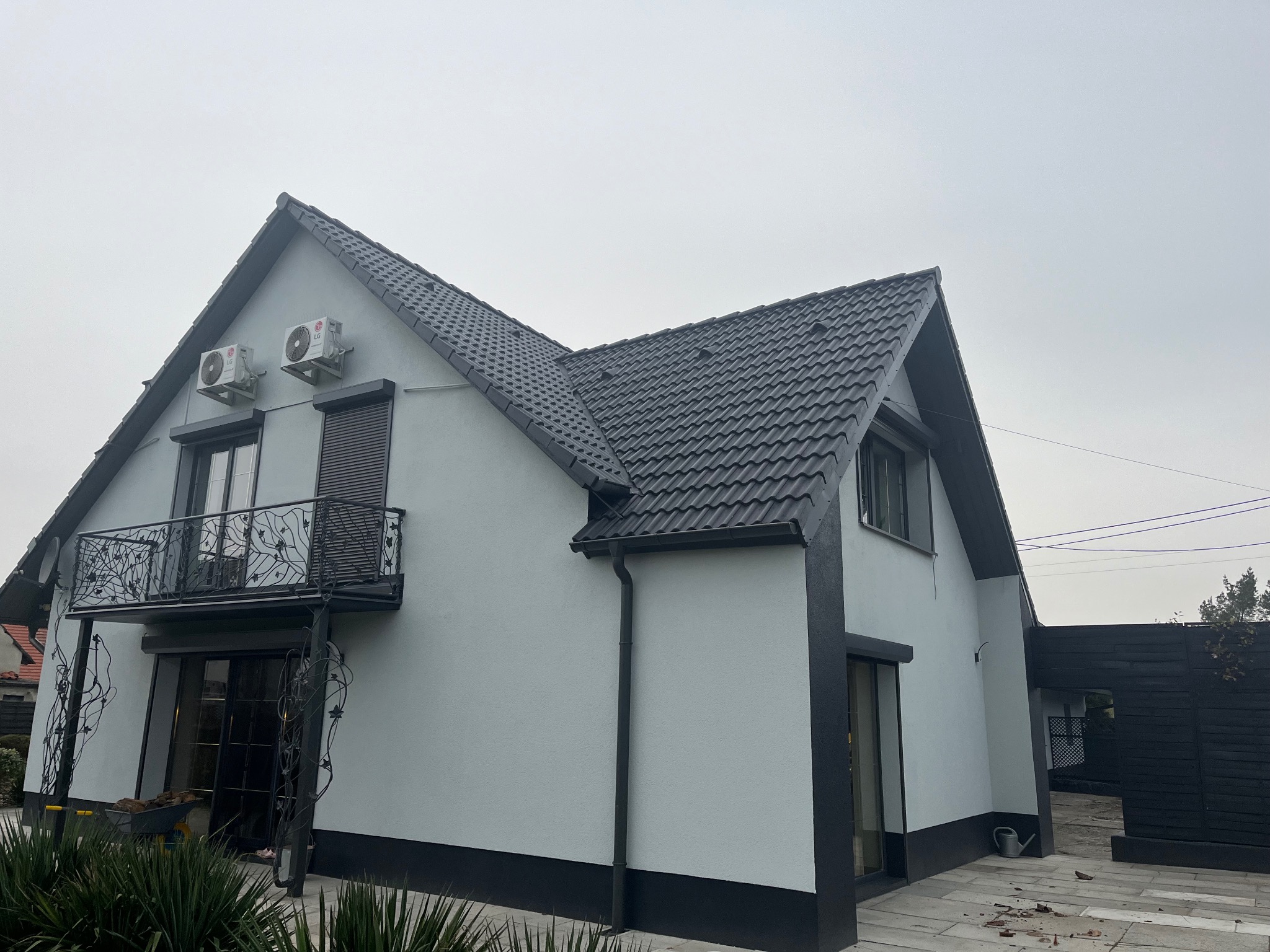 Dom jednorodzinny z ciemną dachówką i jasnoszarą elewacją. Widoczne rynny, okna z roletami i balkon z ozdobną, metalową balustradą. Na elewacji zamontowane klimatyzatory.