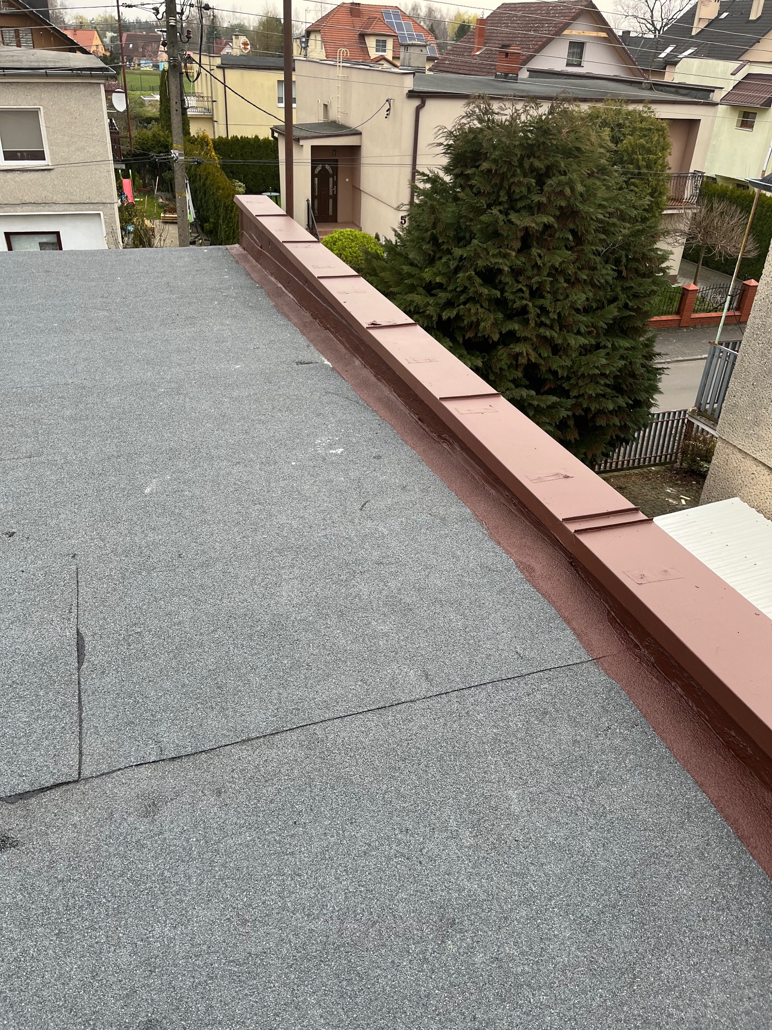 Szary dach pokryty papą z brązową obróbką blacharską i widokiem na zabudowę miejską w tle. Detal wykonania dachu.