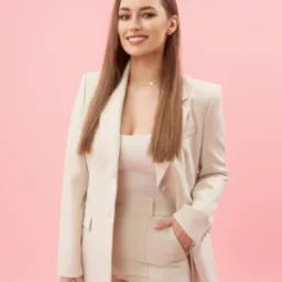 Portret uśmiechniętej kobiety w beżowym garniturze na różowym tle. Długie, proste włosy, delikatna biżuteria. Profesjonalny wizerunek.