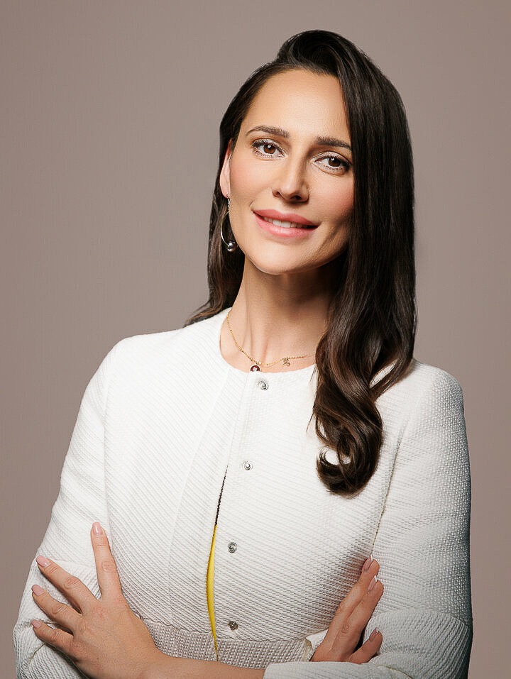 Portret kobiety w białym żakiecie na neutralnym tle. Uśmiechnięta, z rękami skrzyżowanymi, subtelna biżuteria, ciemne włosy. Profesjonalny wizerunek.