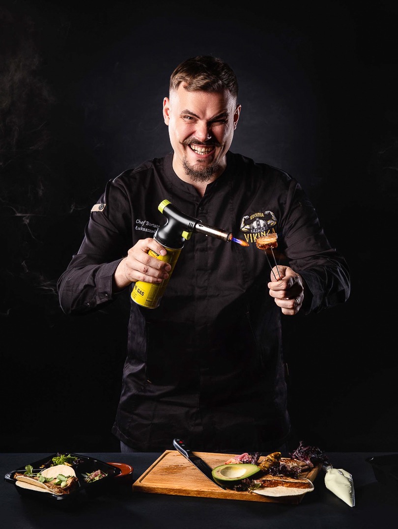 Uśmiechnięty kucharz w czarnym uniformie opala palnikiem danie na desce. W tle czarne tło, a na pierwszym planie gotowe potrawy. Nietypowe ujęcie kulinarne.