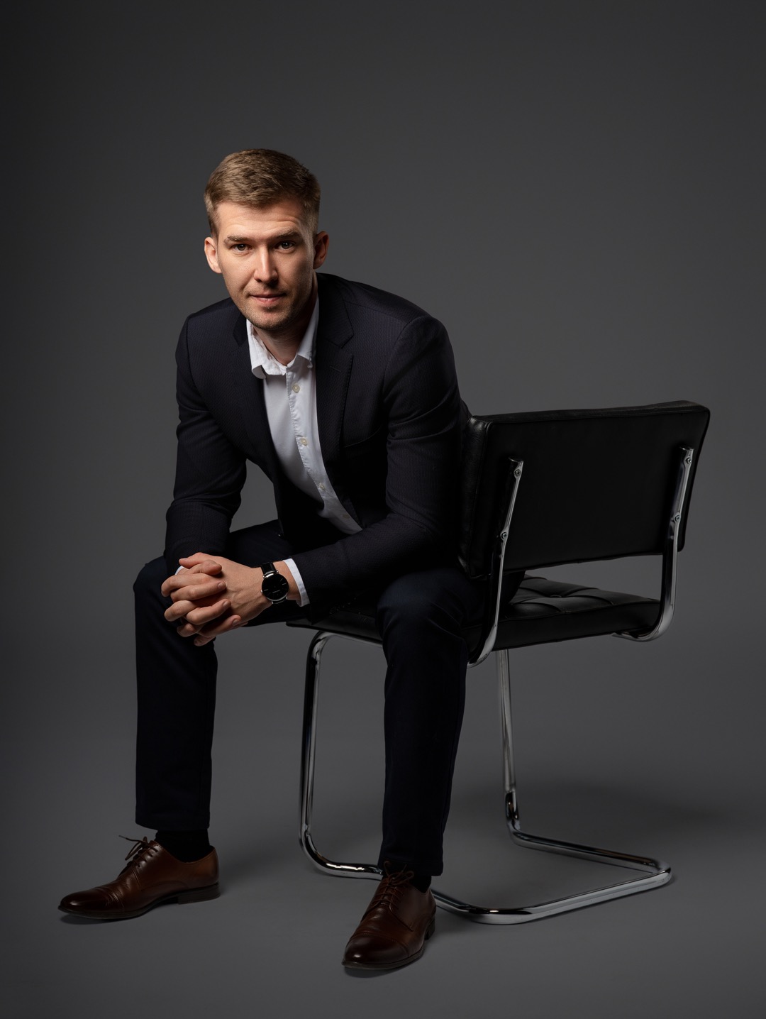 Mężczyzna w garniturze siedzi na krześle z rękami złożonymi na kolanach. Widoczny zegarek i brązowe buty. Szare tło, studyjne oświetlenie.