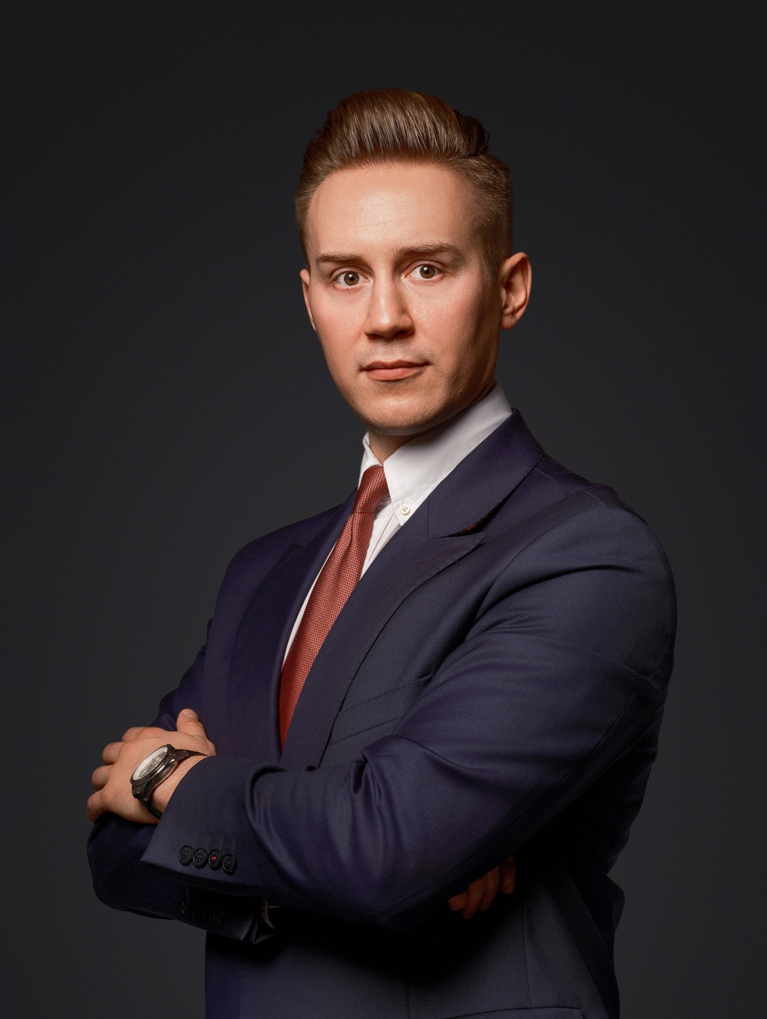 Portret biznesowy mężczyzny w granatowym garniturze i krawacie w odcieniach czerwieni, z zegarkiem na ręce, na ciemnoszarym tle.