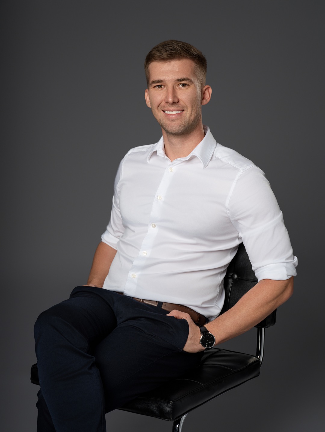 Uśmiechnięty mężczyzna w białej koszuli i ciemnych spodniach siedzi na czarnym krześle na tle szarego tła. Na nadgarstku ma zegarek.