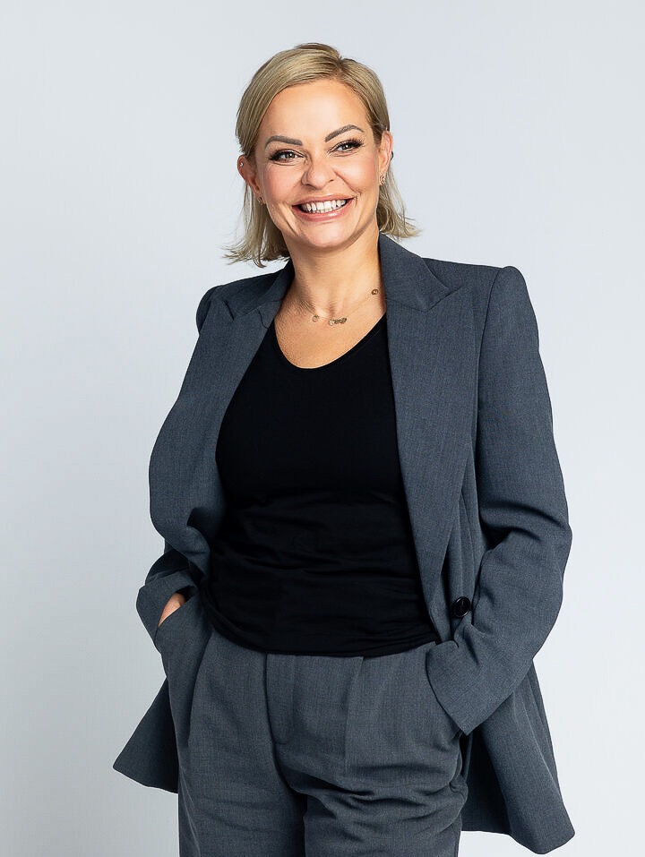 Profesjonalny portret uśmiechniętej blondynki w szarym garniturze na jasnym tle, z rękami w kieszeniach, ukazujący pewność siebie i pozytywne nastawienie.