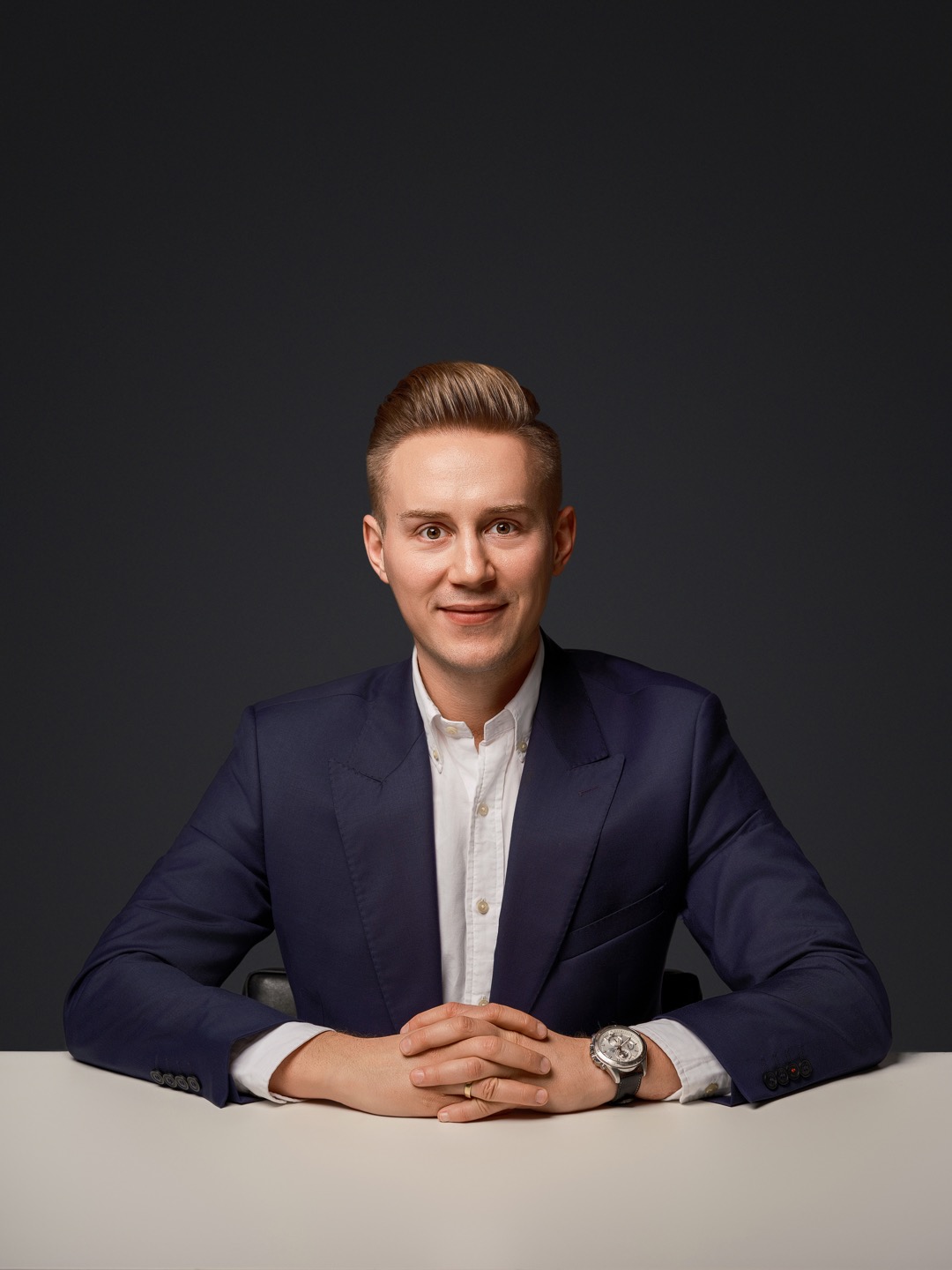 Portret biznesowy mężczyzny w granatowym garniturze, siedzącego za białym stołem na tle ciemnoszarego tła. Widoczny zegarek na lewym nadgarstku.