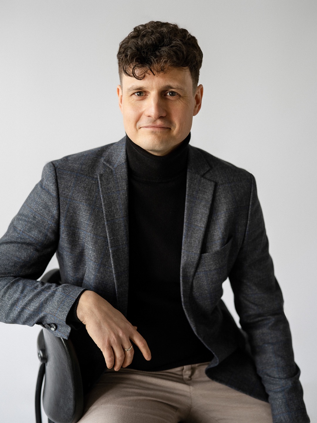 Portret mężczyzny w szarej marynarce i czarnym golfie, siedzącego na krześle na jasnym tle. Uśmiechnięty, z dłonią opartą o oparcie krzesła. Widoczny pierścionek.
