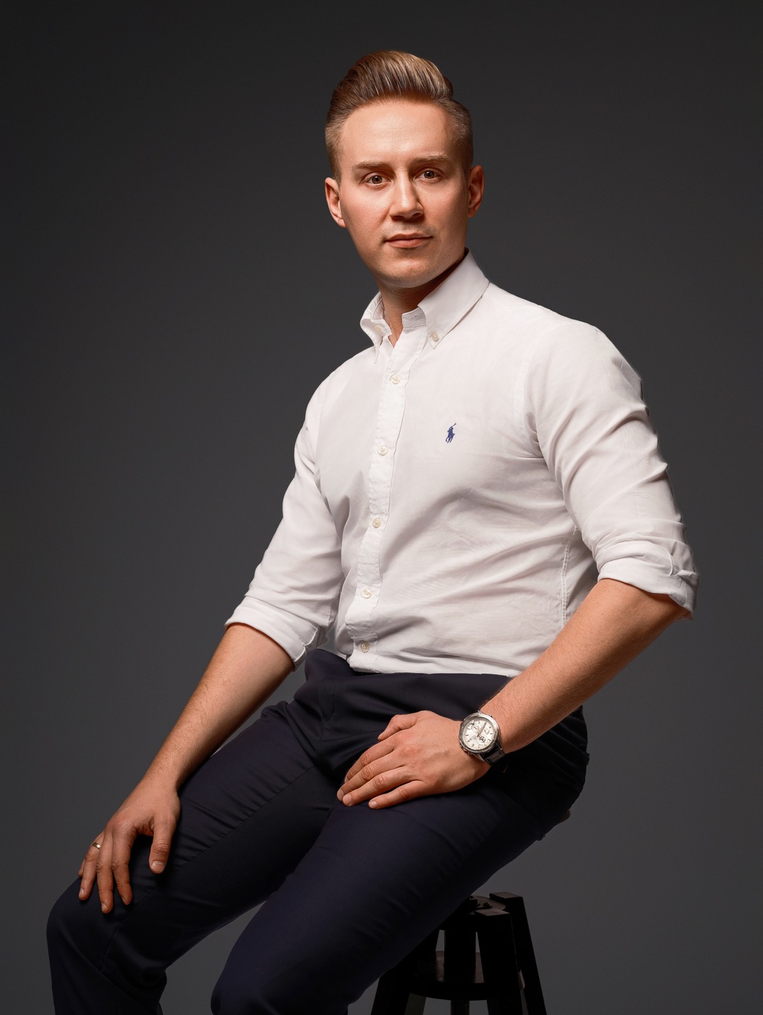 Portret biznesowy młodego mężczyzny w białej koszuli i ciemnych spodniach, siedzącego na stołku na szarym tle. Widoczny zegarek i pierścionek.