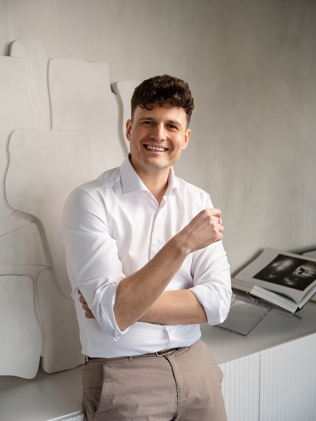 Uśmiechnięty mężczyzna w białej koszuli i beżowych spodniach pozuje na tle minimalistycznej ściany i książki. Sesja biznesowa w neutralnej tonacji.