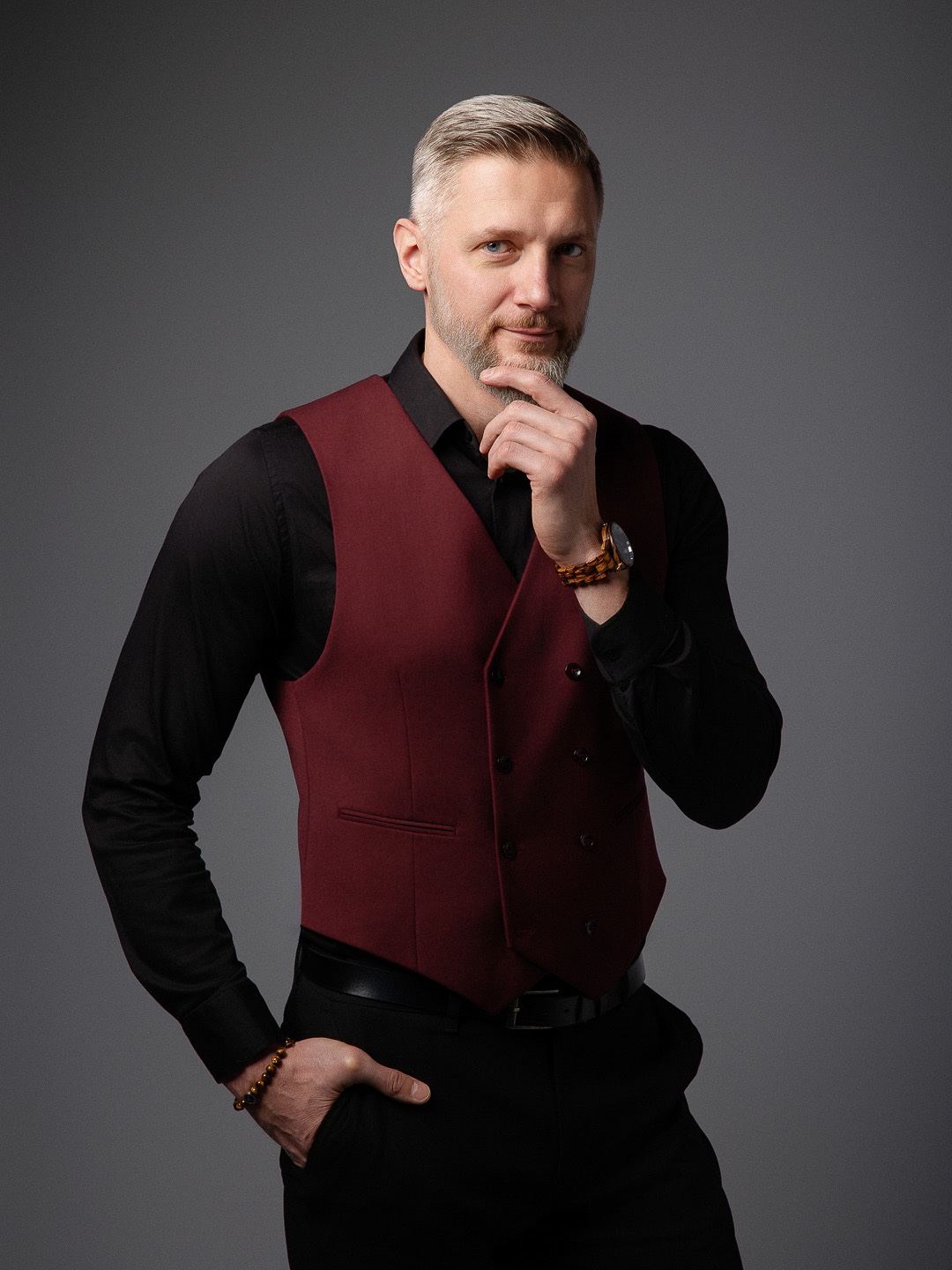 Mężczyzna z brodą w bordowej kamizelce, czarnej koszuli i spodniach pozuje na szarym tle, trzymając rękę na brodzie i w kieszeni, prezentując profesjonalny wizerunek.