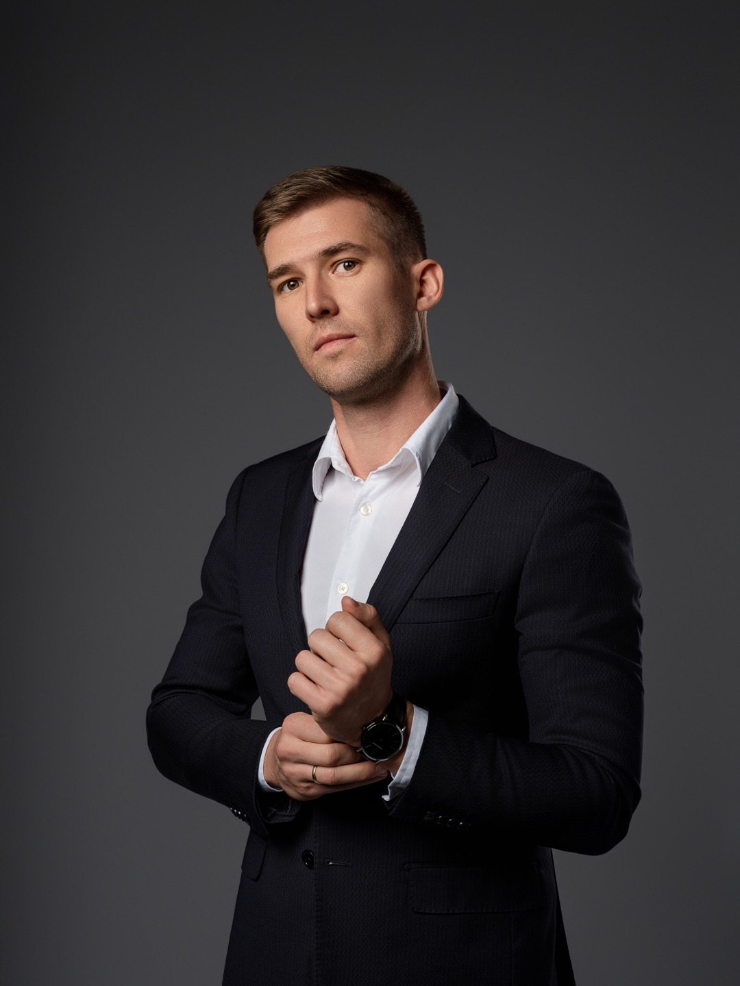 Portret biznesowy mężczyzny w ciemnym garniturze i białej koszuli, zapinającego mankiet zegarka na tle szarego studia.
