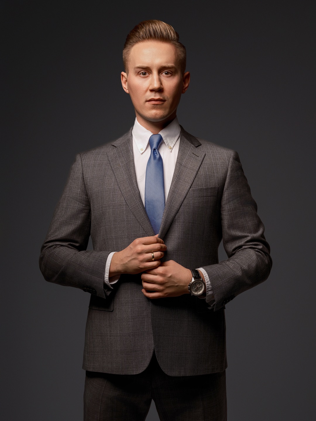 Mężczyzna w szarej kraciastej marynarce, białej koszuli i błękitnym krawacie, zapina marynarkę. Widoczny zegarek na ręce i złoty pierścionek. Stonowane oświetlenie.