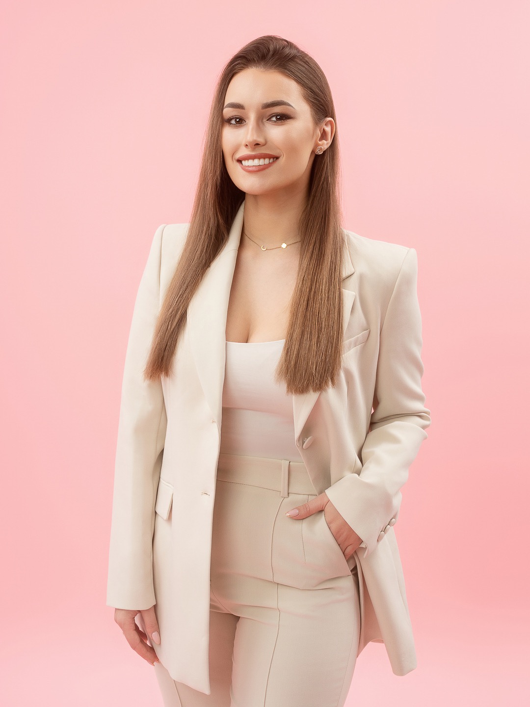 Portret uśmiechniętej kobiety w beżowym garniturze na różowym tle. Długie, proste włosy, delikatna biżuteria. Profesjonalny wizerunek.