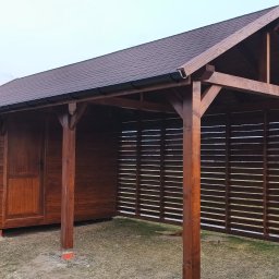 BERWOOD Jarosław Chwasta - Drewniana altana z zadaszeniem z gontu bitumicznego, z ażurową ścianą boczną. Widoczne elementy konstrukcyjne i wykończeniowe w ciepłym odcieniu brązu.