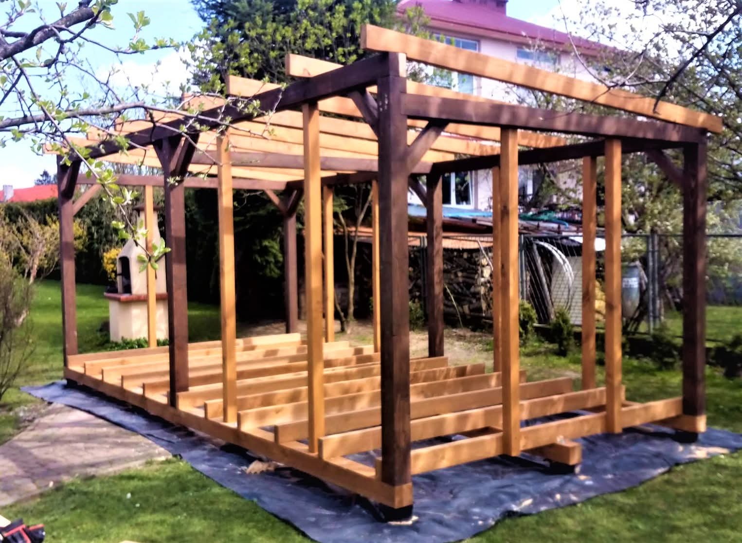 Wznoszona drewniana pergola z ciemnymi słupami i jasnymi belkami na trawniku, widoczny fundament i konstrukcja dachu, w tle dom i zieleń.
