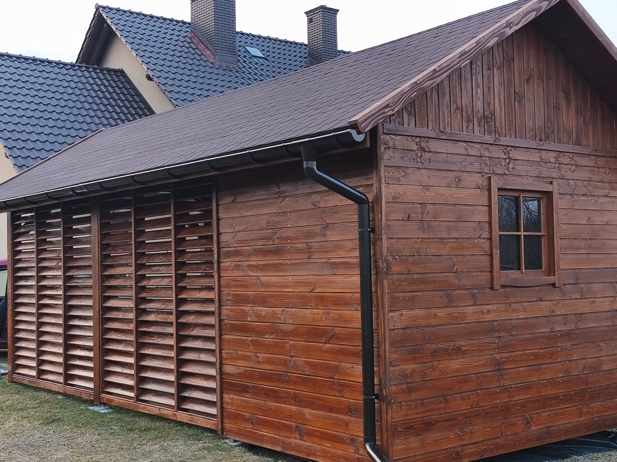 Drewniana altana z brązowym dachem i żaluzjowymi ścianami, widoczna z boku na tle domu z ciemną dachówką. Detal: czarna rynna.
