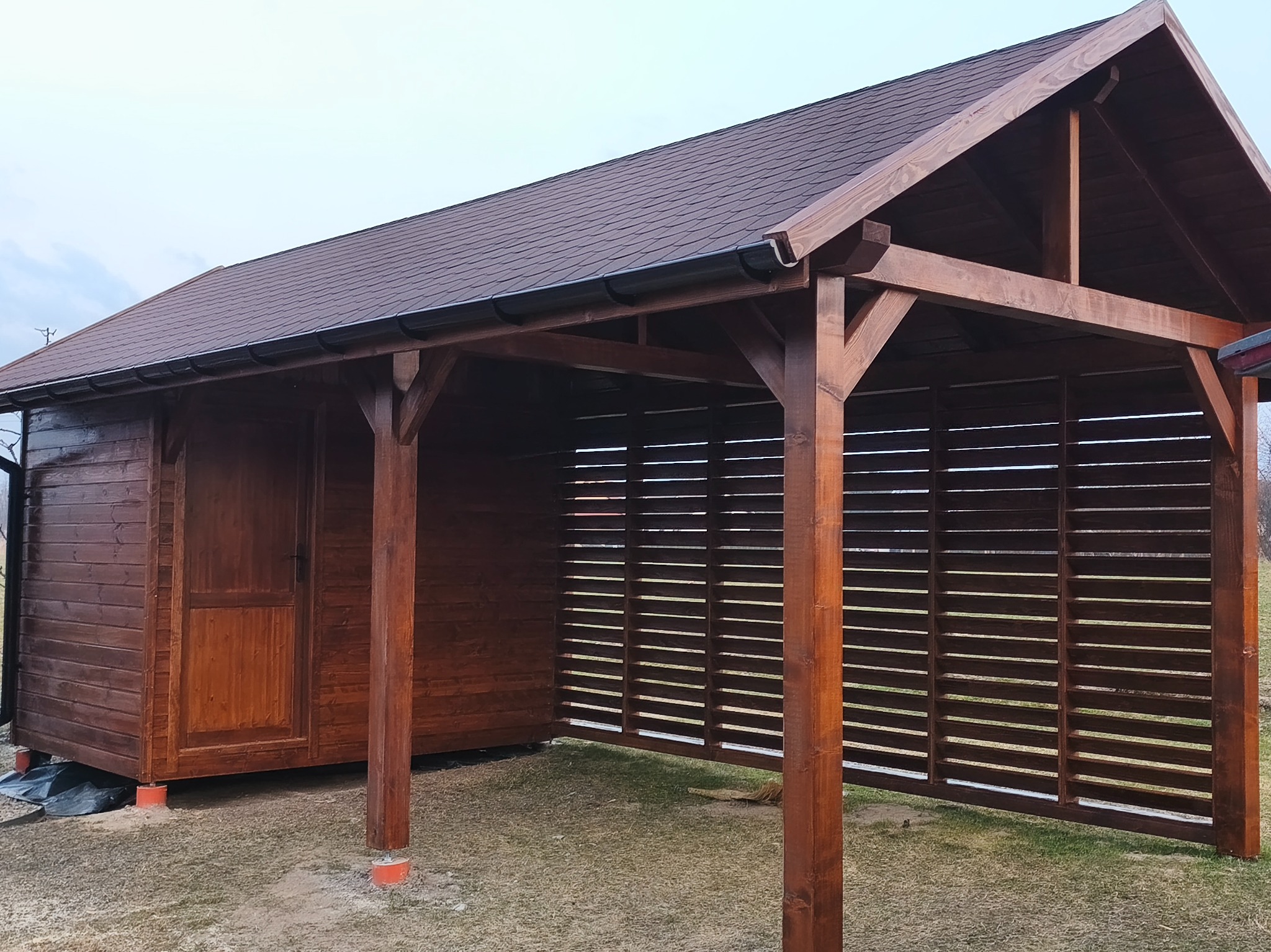 Drewniana altana z zadaszeniem z gontu bitumicznego, z ażurową ścianą boczną. Widoczne elementy konstrukcyjne i wykończeniowe w ciepłym odcieniu brązu.